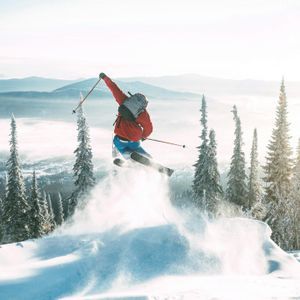 A skier in a red jacket and backpack jumps mid-air, kicking up powder snow on a mountain surrounded by snow-covered trees.