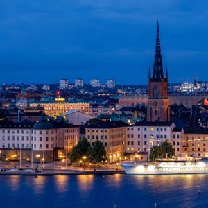 Un panorama urbano illuminato con un'imponente guglia di cattedrale, visto dall'altra parte dell'acqua al tramonto, con una nave attraccata lungo il lungomare.
