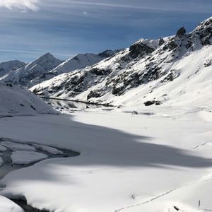 Un lago parzialmente ghiacciato si snoda attraverso una valle innevata circondata da cime montuose illuminate dal sole sotto un cielo blu.