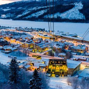 Una vista dall'alto di un villaggio innevato illuminato al crepuscolo, con un lago ghiacciato e montagne sullo sfondo.