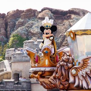 Mickey Mouse, vêtu d'un costume royal blanc et or, salue depuis un char de parade orné, avec un château et des montagnes en arrière-plan.