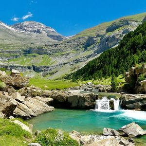 Un río turquesa con una pequeña cascada fluye a través de un valle de montaña verde bajo un cielo azul claro.