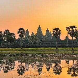 An ancient temple with tall spires and palm trees reflected in a calm body of water against an orange sky at sunrise.