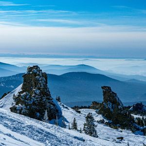 Formazioni rocciose frastagliate sporgono da un fianco di montagna innevato, affacciandosi su strati di montagne azzurre e sfumate che si estendono all'orizzonte sotto un cielo luminoso.