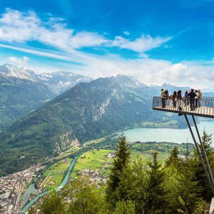 Un gruppo WeRoad ammira una vista panoramica mozzafiato su una valle montana, un lago e una città da una piattaforma sospesa.