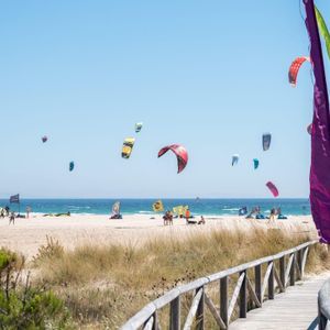 Una pasarela de madera conduce a una playa soleada con muchas cometas de kitesurf coloridas en el cielo sobre el océano.