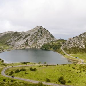Un lago de montaña, rodeado de verdes colinas y picos rocosos, con carreteras para vehículos que serpentean a lo largo de su orilla.