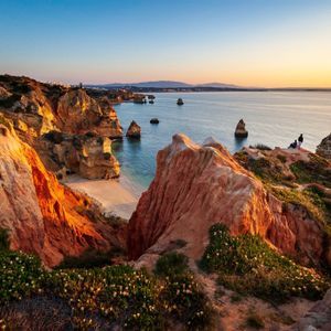 Rugged red cliffs covered in green vegetation overlook a calm sea with sea stacks and a sandy cove at sunset.