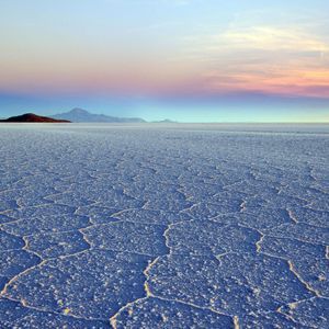Una vasta pianura salata con motivi esagonali si estende verso montagne distanti sotto un cielo colorato all'alba.