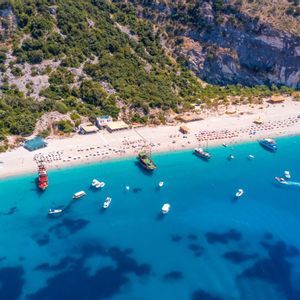 Una vista aérea de una playa de arena concurrida con muchos barcos flotando en el agua turquesa brillante al pie de una ladera verde.