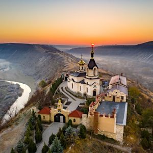 Un monastero ortodosso con cupole dorate sorge su un crinale che domina una valle fluviale nebbiosa all'alba.