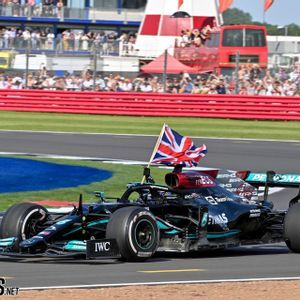 A black Formula 1 car with a British flag drives on a racetrack as spectators watch from the grandstands in the background.