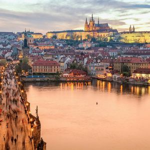 Una fotografía de larga exposición de un concurrido puente histórico sobre un río al atardecer, con un paisaje urbano iluminado y un castillo de fondo.