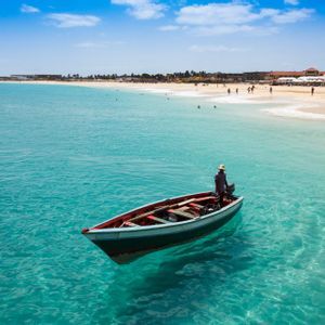Une personne coiffée d'un chapeau se tient dans un petit bateau sur une eau turquoise et claire, avec une plage de sable et des gens à l'arrière-plan.