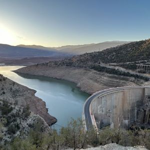 Una grande diga in cemento con una strada in cima si curva attraverso un bacino idrico in una valle montuosa all'alba.