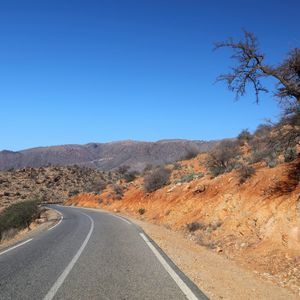 Una strada asfaltata si snoda attraverso un paesaggio arido e collinare con terra rossa e vegetazione rada sotto un cielo blu chiaro.