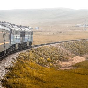 Un lungo treno passeggeri con una locomotiva blu viaggia su un binario curvo attraverso una vasta pianura erbosa con colline nella distanza nebbiosa.