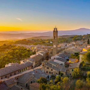 Una vista aerea di una città storica con una torre dell'orologio, circondata da verdi colline e montagne al tramonto.