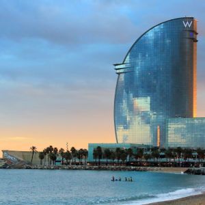 Un moderno hotel di vetro a forma di vela si erge vicino a una spiaggia sabbiosa al tramonto, con palme e persone che fanno paddleboard in mare.