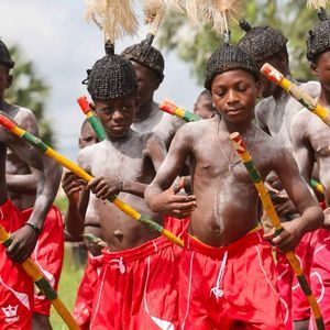 Un gruppo di giovani ragazzi in abiti tradizionali con capelli intrecciati e pittura corporale tiene bastoni colorati durante una cerimonia.
