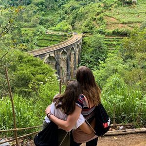 Dos mujeres de un viaje en grupo de WeRoad se abrazan mientras miran un puente de arco de piedra en un valle exuberante y verde.