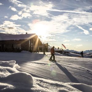 Ein Skifahrer auf einer schneebedeckten Piste vor einer Berghütte, wo die tief stehende Sonne lange Schatten erzeugt.