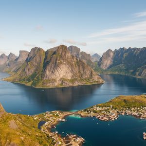 Luftaufnahme eines Küstendorfs am Fuße großer, felsiger Berge, umgeben von einem tiefblauen Fjord.