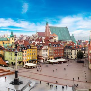 Una veduta dall'alto di una storica piazza cittadina con edifici colorati, un'alta colonna e una torre dell'orologio, sotto un cielo azzurro e luminoso.
