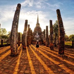 Una persona si erge tra antichi pilastri di fronte a una statua di Buddha e a uno stupa, con lunghe ombre proiettate dal sole al tramonto.