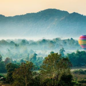 Una mongolfiera colorata fluttua su una valle nebbiosa e verde con montagne sullo sfondo all'alba.