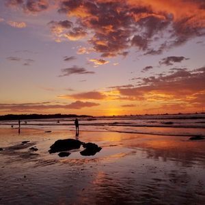 Des silhouettes de personnes sur une plage mouillée à marée basse, avec un coucher de soleil aux couleurs orange et violet qui se reflète sur le sable et l'eau.