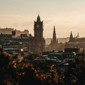 Uno storico skyline cittadino con una grande torre dell'orologio e guglie, stagliato contro un cielo caldo e velato al tramonto.
