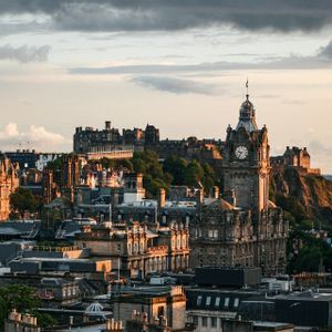 A historic city skyline with a castle on a hill and a prominent clock tower, bathed in the warm light of sunset.