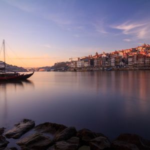 Una barca de madera tradicional en un río tranquilo al atardecer, con los coloridos edificios de una ciudad en la ladera en la orilla opuesta, bajo un cielo rosado y azul.