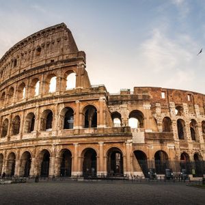 El antiguo anfiteatro Coliseo en Roma, visto desde una plaza de adoquines vacía al amanecer bajo un cielo parcialmente nublado.