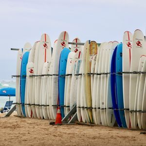 Una grande rastrelliera metallica contiene numerose tavole da surf in posizione verticale su una spiaggia sabbiosa, con l'oceano sullo sfondo.
