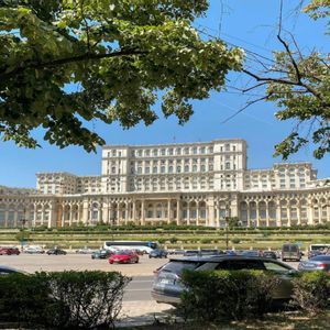 Un maestoso e sfarzoso palazzo, visibile da una strada e un parcheggio, incorniciato da alberi verdi e frondosi sotto un cielo azzurro e limpido.
