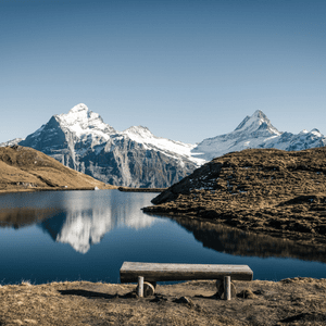 An empty wooden bench on a grassy shore overlooks a calm lake reflecting snow-capped mountains under a clear sky.