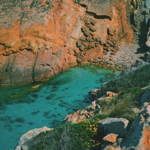 Vista dall'alto di una caletta rocciosa con acqua cristallina turchese, circondata da ripide scogliere rosso-arancio e vegetazione verde.