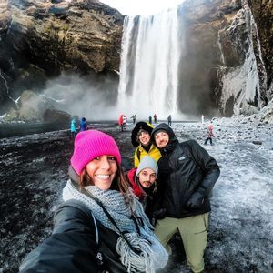A WeRoad group trip of four people takes a selfie in front of a large waterfall on a snowy and icy shore.