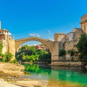 Un ponte ad arco in pietra attraversa un fiume turchese, fiancheggiato da edifici storici e una vegetazione lussureggiante, sullo sfondo di un cielo azzurro luminoso.