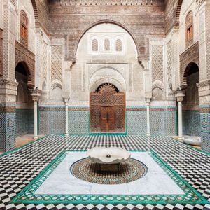 Un patio ornamentado con una fuente central sobre un suelo de baldosas a cuadros blancas y negras, rodeado de paredes con intrincados azulejos y arcos tallados.