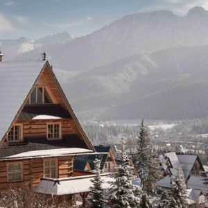 Un chalet de madera con tejado nevado domina un pueblo de montaña y pinos, con grandes picos nevados de fondo.