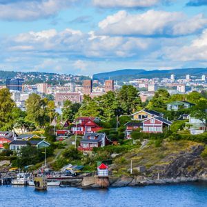 Colorful houses dot a green, rocky shoreline in front of a distant city skyline under a partly cloudy sky.