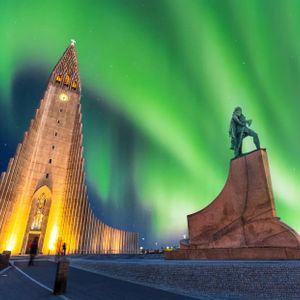 A modern church and a statue stand illuminated at night under the vibrant green Northern Lights.