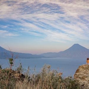 Una persona con un cappello è seduta su una grande roccia, ammirando un vasto lago e montagne lontane sotto un cielo azzurro nuvoloso.