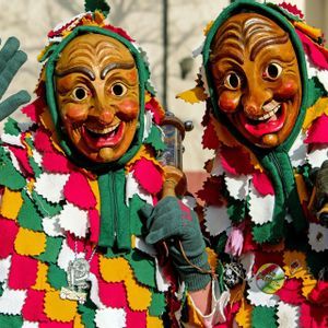 Due persone in costumi colorati a patchwork e maschere di legno sorridenti posano a un carnevale, una delle quali saluta.