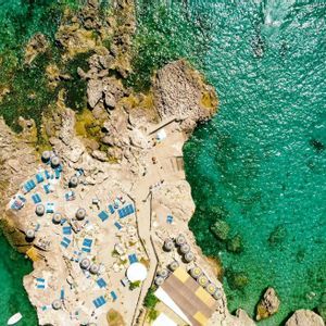 An aerial top-down view of a rocky coastline with blue sun loungers and umbrellas next to vibrant turquoise water.