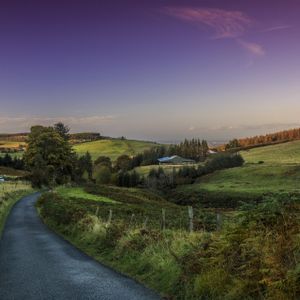 Eine befestigte Landstraße schlängelt sich durch sanfte, grüne Hügel, gesprenkelt mit Bauernhöfen, unter einem farbenprächtigen Sonnenuntergangshimmel.
