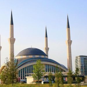 Una grande moschea moderna con una cupola blu e quattro alti minareti si erge in un parco erboso sotto un cielo limpido.